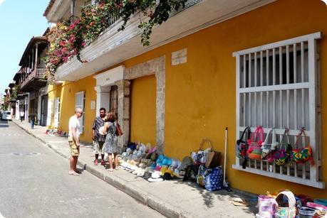 vente de souvenirs (chapeaux, sacs) dans une rue colorée de Carthagène, un couple discute avec un vendeur